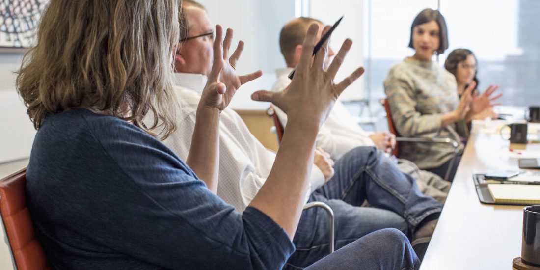 office workers sitting along side of long table- women in foreground with hands gesturing