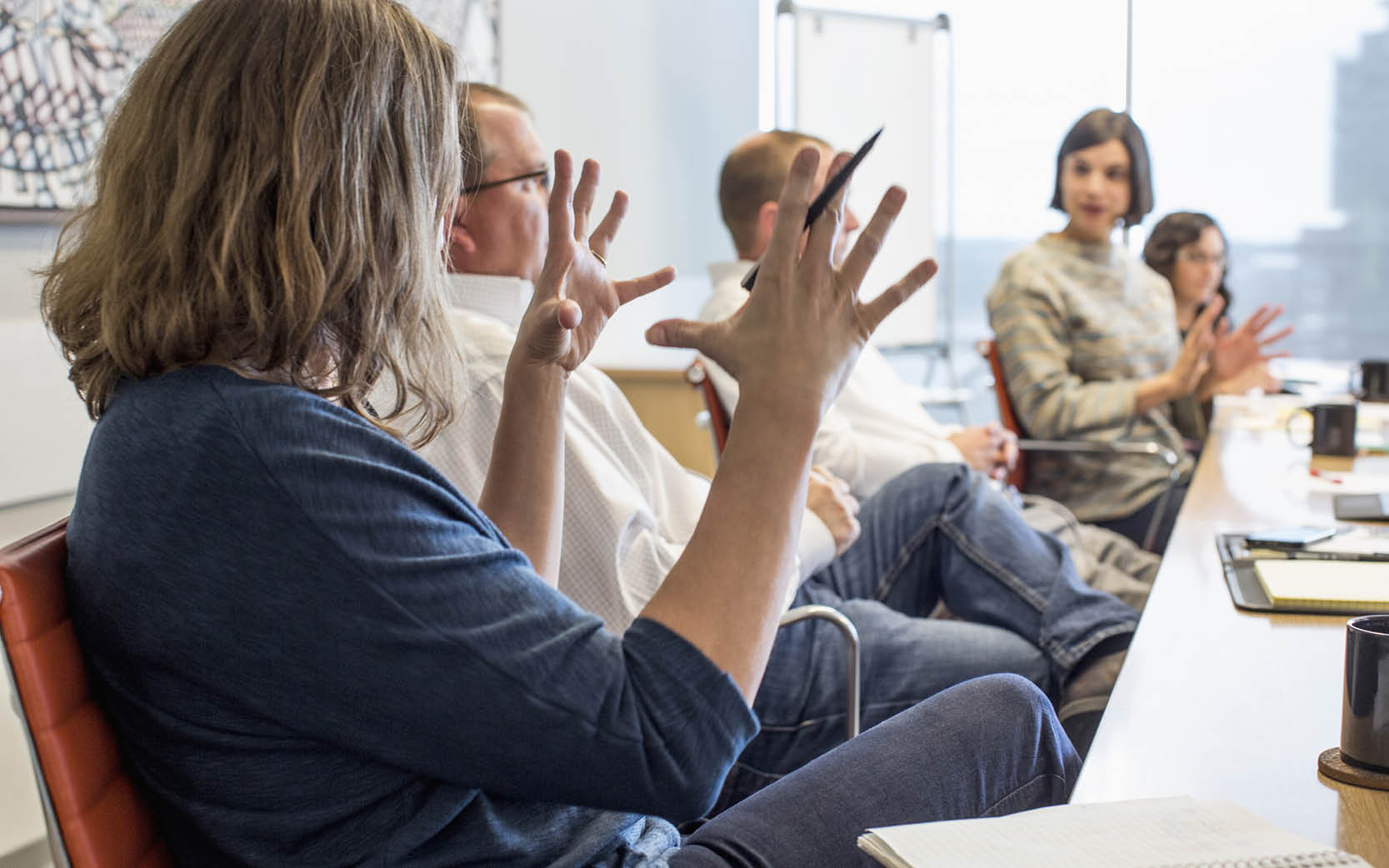 office workers sitting along side of long table- women in foreground with hands gesturing