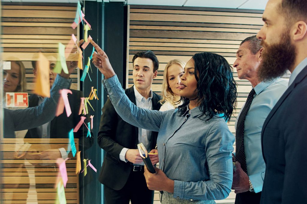 office workers standing meeting pointing to post it notes on wall